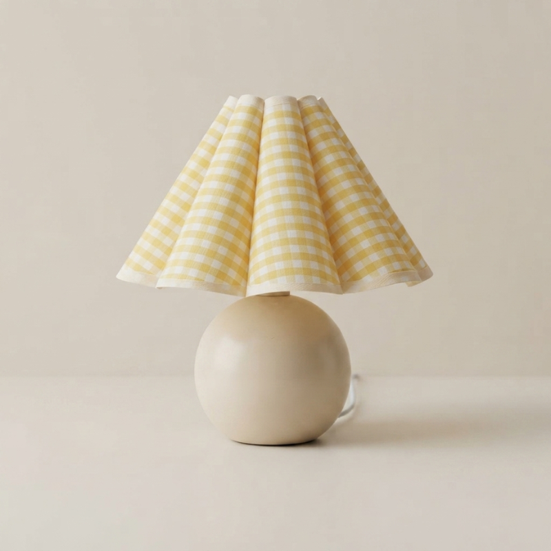 A table lamp with a pleated yellow and white gingham fabric shade and a smooth, matte cream-colored spherical base, set against a neutral beige background.