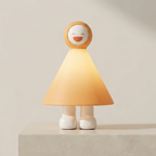 A whimsical child-friendly table lamp shaped like a smiling cartoon girl, standing on a light stone pedestal against a warm beige background. The lamp features a glowing warm yellow-orange cone shade, a round head with a white face and orange smile, and small white legs.