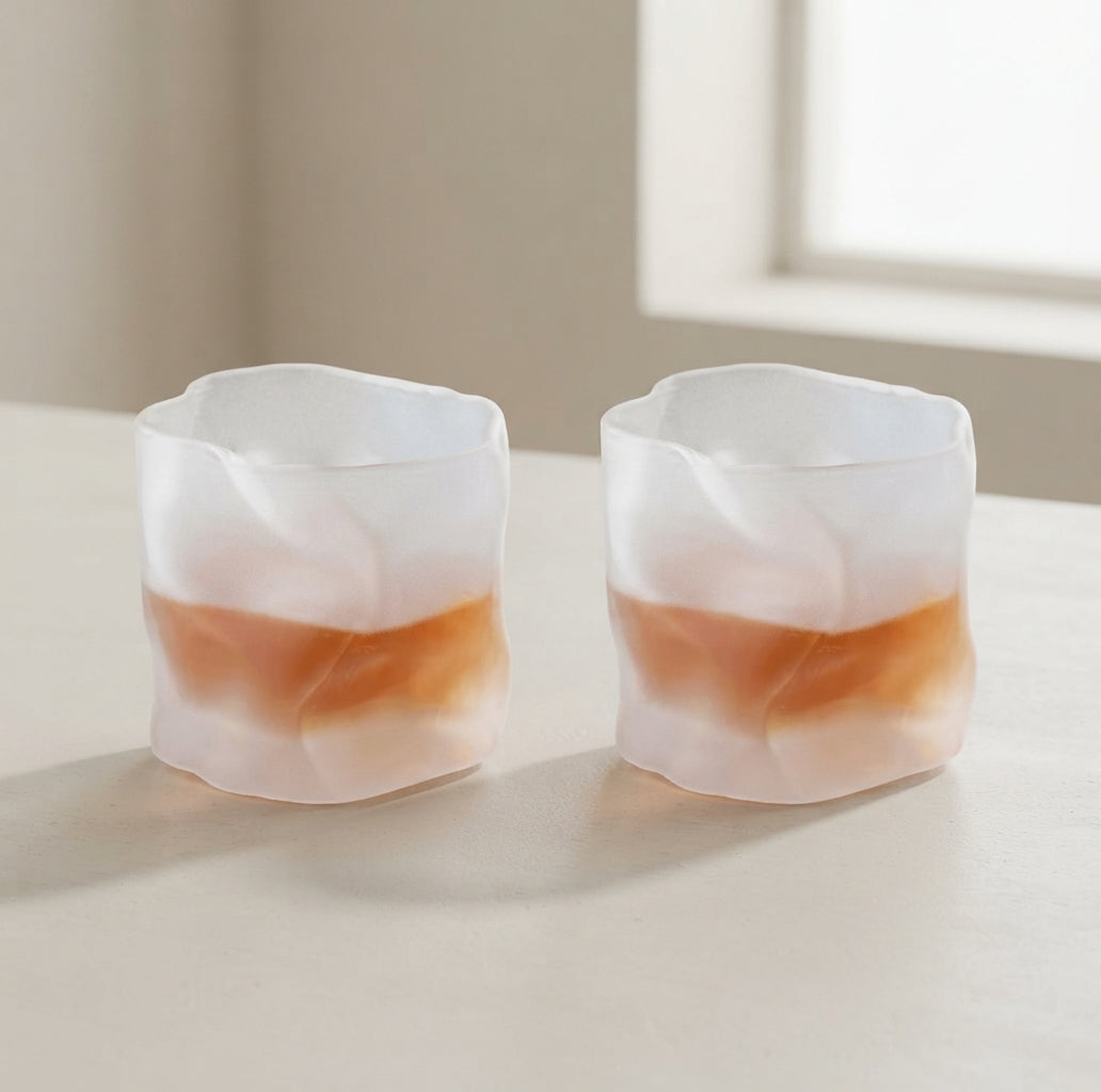 Two small frosted glass cups with a wavy organic shape, filled with an amber-colored liquid, sitting on a light beige tabletop in a softly lit room.
