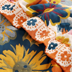 A close-up of a colorful floral quilted fabric, showing a ruffled border in orange, white, and blue layered over a larger floral pattern in yellow, blue, and gray.