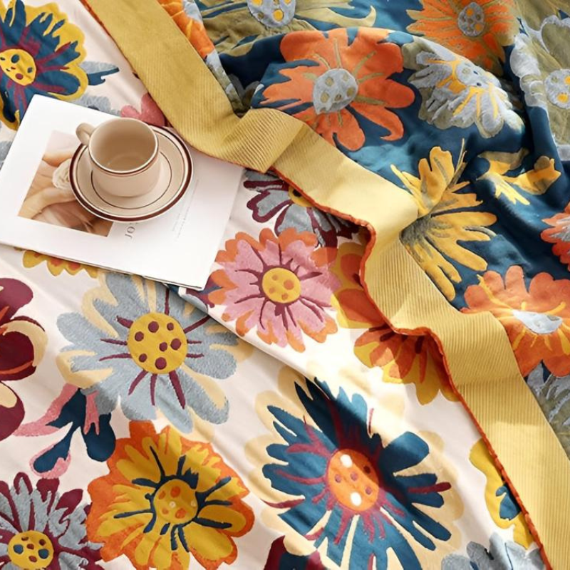 A close-up of a colorful floral blanket or throw with large daisy-like flowers in blue, orange, yellow, pink, and white, draped across a bed-like surface with a folded cup and saucer and a small card placed on top.