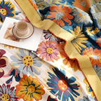 A close-up of a colorful floral blanket or throw with large daisy-like flowers in blue, orange, yellow, pink, and white, draped across a bed-like surface with a folded cup and saucer and a small card placed on top.