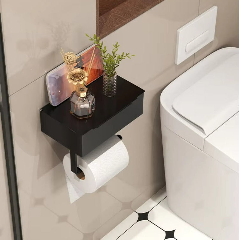 A black wall-mounted toilet paper holder shelf mounted on a beige tiled bathroom wall next to a white toilet. The shelf holds a small potted green plant and a decorative gold object, with a roll of white toilet paper hanging below on a black bracket.