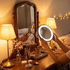 Person using a round light-up mirror next to a larger vanity mirror with decorative items on a table.
