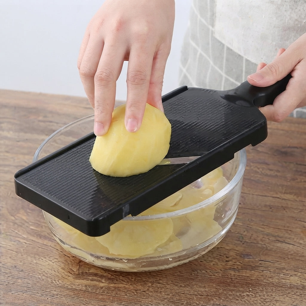 A black multi-functional vegetable slicer and grater with a handle is being used by a person to slice a peeled potato over a glass bowl on a wooden table.