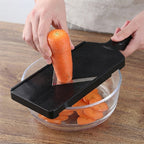 A black rectangular vegetable slicer with a triangular blade is being used by two hands to slice a carrot over a clear glass bowl on a wooden table.