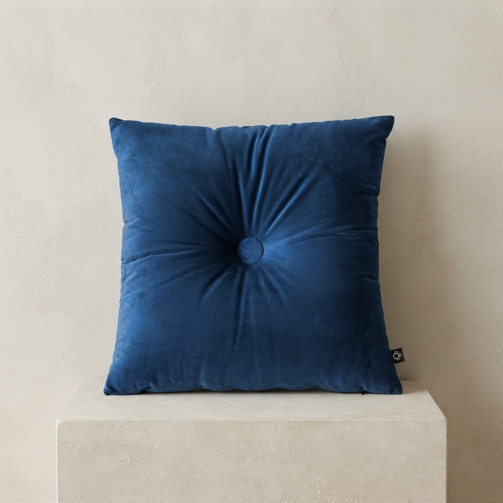 A square navy blue velvet throw pillow with a central button tuft, resting on a light beige stone pedestal against a textured beige wall.