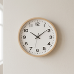 A minimalist analog wall clock with a light natural wood frame, white face, and black numerals and hands, mounted on a warm off-white wall.