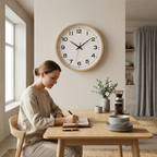 A woman sits at a wooden dining table writing in a notebook in a bright, modern room. A large round wall clock with a light wood frame and white face hangs on the wall behind her.