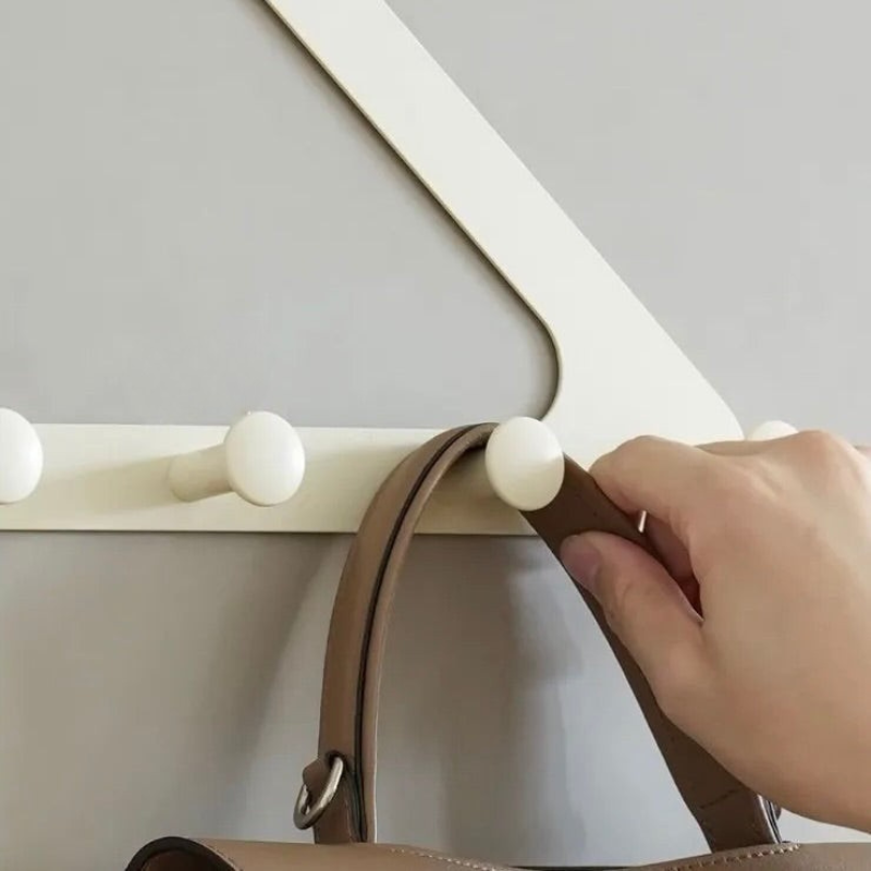 A close-up of a hand hanging a brown handbag strap from a white wall-mounted hook rack against a light gray wall.
