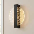 A modern wall sconce light fixture with a vertical black rectangular frame and a circular glass shade filled with gold glitter, illuminated against a beige wall.