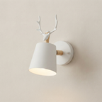 A modern wall-mounted light fixture shaped like a white deer head with antlers above a tapered white lampshade, with light wood accents and a round white wall plate, shown against a warm beige wall.