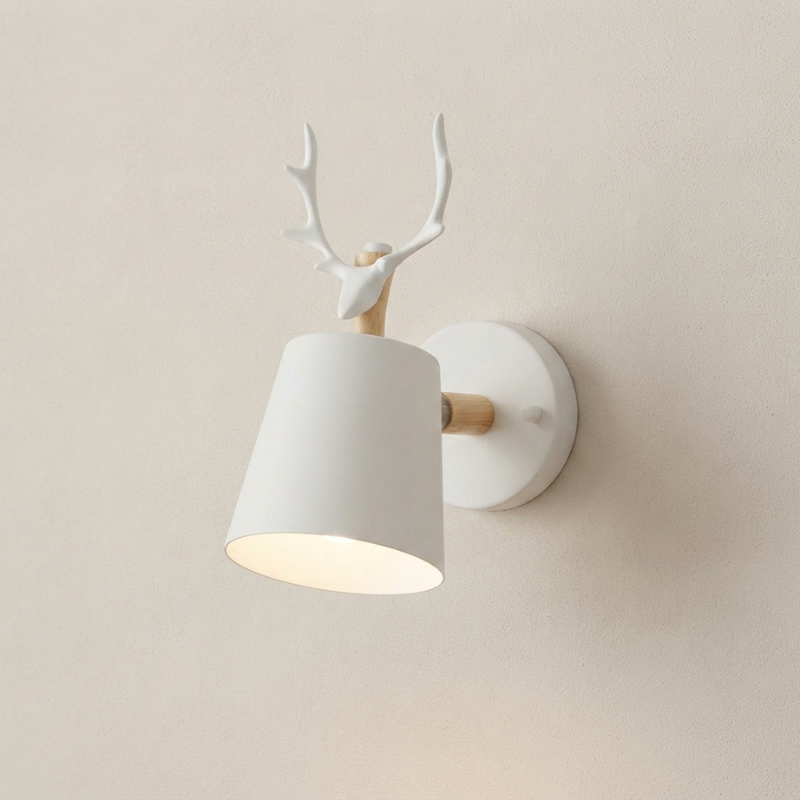 A modern wall-mounted light fixture shaped like a white deer head with antlers above a tapered white lampshade, with light wood accents and a round white wall plate, shown against a warm beige wall.