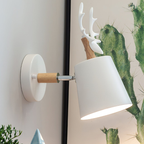 A modern wall-mounted lamp with a white conical shade, a light wood cylindrical stem, and a white antler-like top, shown attached to a white wall next to a framed print featuring green cacti.
