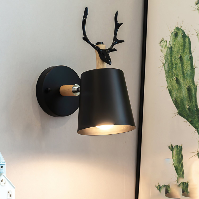 A modern wall-mounted light fixture shaped like a black deer head with antlers above a tapered black lampshade, shown lit against a light wall next to a framed cactus print.