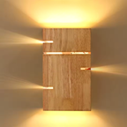 A modern rectangular wall sconce with a light natural wood panel and multiple horizontal cutouts emitting warm yellow light upwards and downwards, set against a neutral beige wall.
