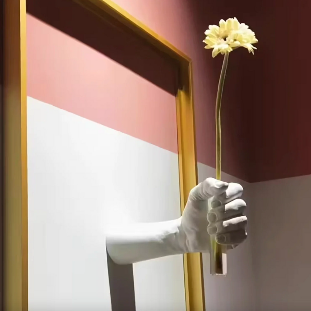 Hand in a white glove holding a yellow flower against a colorful wall.