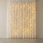 Warm white curtain string lights glowing through a white sheer curtain panel hanging in front of a neutral beige wall.