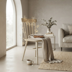 A light natural wood spindle-back chair in a softly styled living room, draped with a beige throw blanket and a small book, with warm fairy lights glowing behind it. A sofa, side table, and woven rug are visible in the background.