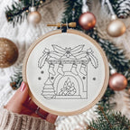 A hand holds a wooden embroidery hoop containing black line art of a holiday fireplace with stockings, holly, a tree, and snowflakes on white fabric. The background shows a softly blurred Christmas tree with green branches and metallic ornaments.