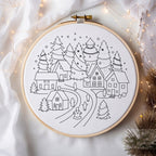 A circular embroidery hoop containing a black line-art winter village design on white fabric, styled with draped white fabric, warm fairy lights, and decorative pine branches around it.