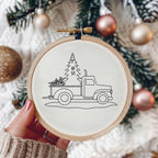 A hand with pink nail polish holds a wooden embroidery hoop featuring a black line art design of a vintage pickup truck carrying a Christmas tree. The background is a soft-focus holiday scene with pine branches and metallic rose gold and gold Christmas ornaments.