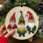 A hand holds a wooden embroidery hoop featuring three festive embroidered gnomes with red, green, white, and brown details on cream fabric, set against a holiday background of pine branches and pinecones.
