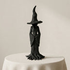 A black, textured figurine of a witch with a tall pointed hat and draped robe, standing on a draped white table against a warm beige background.