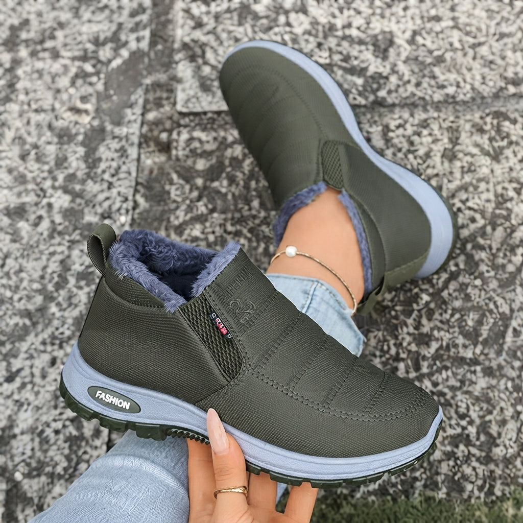 A pair of women's slip-on winter sneakers in olive green with a gray sole and a blue-gray faux-fur lining, shown being worn outdoors on a gray stone pavement.