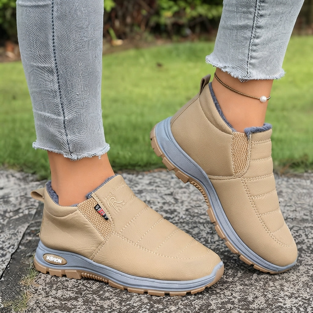 A person wearing beige slip-on winter boots with gray soles, standing on a stone path with green grass in the background.