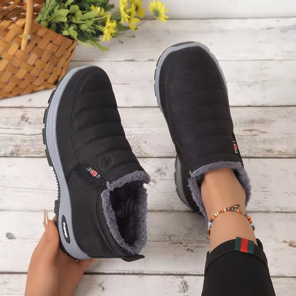 A pair of black quilted winter slip-on shoes with gray soles and a gray fleece lining, shown being worn with one hand holding one shoe on a white wood plank surface.
