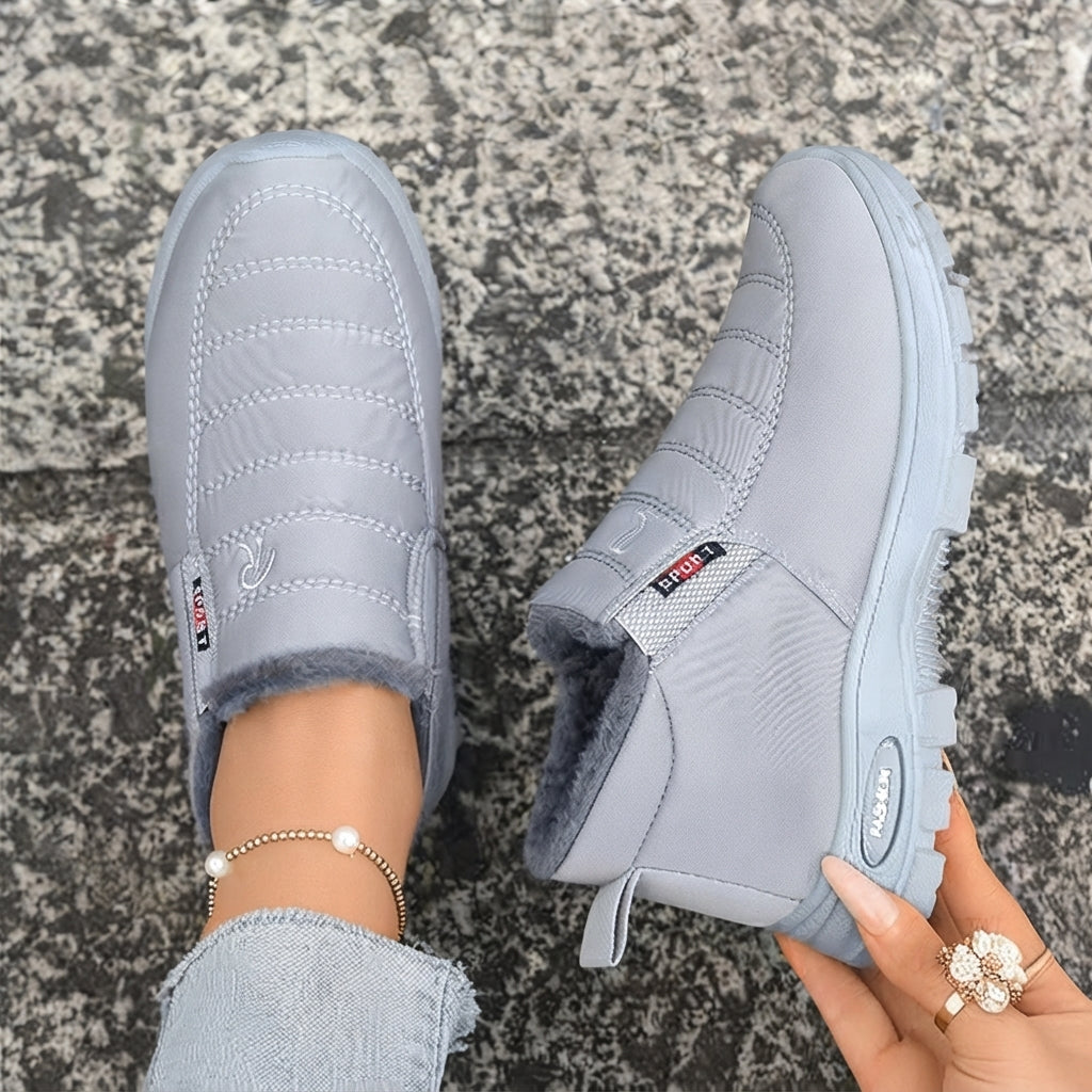 A pair of light gray slip-on winter boots with a fuzzy lining, shown on a person's feet and held by a hand over a gray stone pavement background.