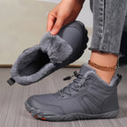 A person wearing gray winter boots with fluffy gray lining, holding the other boot to show the interior. The person is wearing gray jeans and a gold chain bracelet, standing on a gray wood floor.