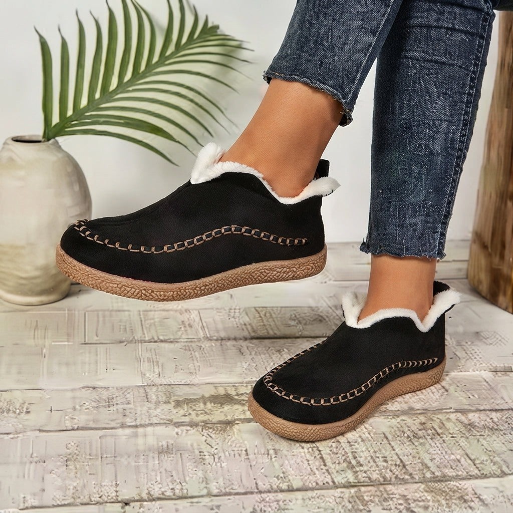 A person wearing black slip-on slippers with white faux-fur trim and braided jute soles, shown with blue jeans in a styled indoor setting with a plant and ceramic vase.