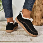 A pair of women's black slip-on moccasin-style slippers with white faux-fur trim and a tan sole, shown on a person wearing blue jeans in a rustic indoor setting.