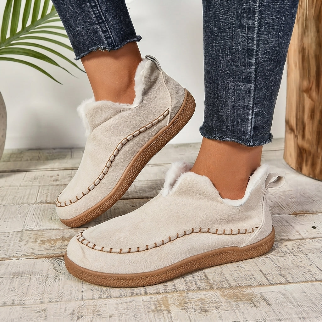 A person wearing a pair of beige ankle-height slip-on shoes with faux fur trim and brown soles, paired with dark blue jeans, standing on a rustic white wooden floor.
