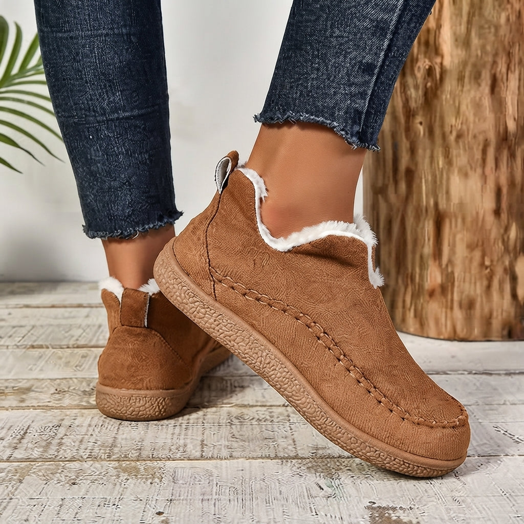 A person wearing brown suede slip-on boots with white faux-fur trim, paired with dark blue frayed-hem jeans, standing on a rustic white wood floor next to a wooden log.