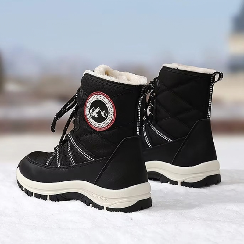 A pair of black winter boots with white faux-fur lining and a circular mountain logo patch on the side, standing on snow with a blurred outdoor background.
