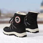 A pair of black winter boots with white faux-fur lining and a circular mountain logo patch on the side, standing on snow with a blurred outdoor background.