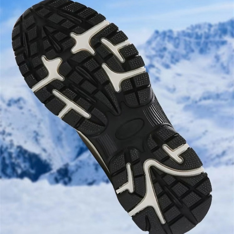 A close-up of a black and white winter hiking boot sole with aggressive tread, set against a blurred snowy mountain background.