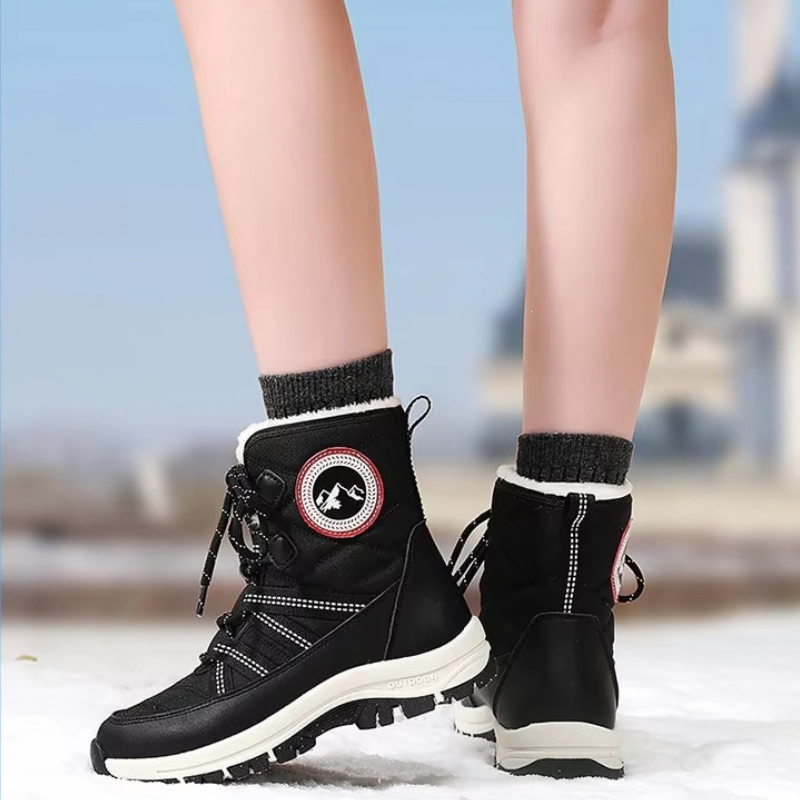 A pair of black mid-calf winter boots with white soles and black socks, shown on a person's feet standing in snow with a blurred outdoor background.