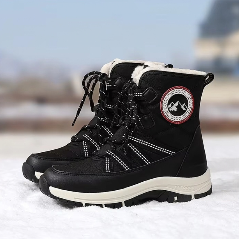 A pair of black winter lace-up boots with white faux-fur lining, white stitching, and a circular mountain logo patch on the side, standing in snow with a blurred outdoor background.