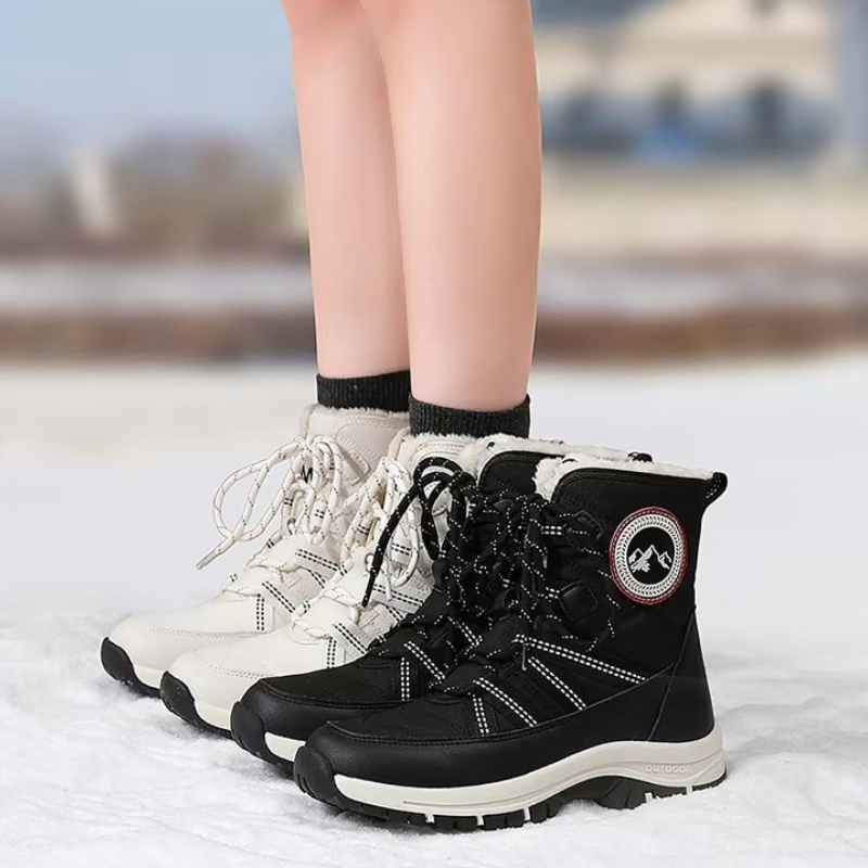 A pair of women's mid-calf winter boots shown on a person's feet in a snowy outdoor setting. The boots are black and off-white, with black laces and white stitching, and feature a circular logo patch on the side.