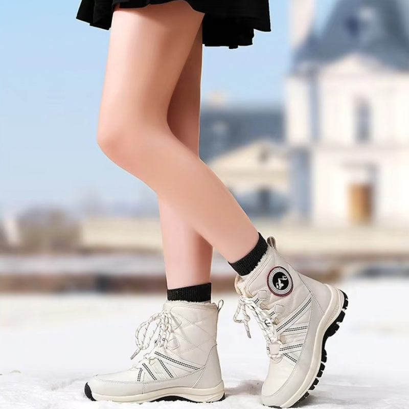A person wearing off-white mid-calf winter boots with black socks, standing outdoors in a snowy setting with a blurred building in the background.