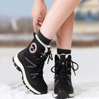 A person wearing black winter lace-up boots with white trim and black socks, standing in snow with a blurred outdoor background.