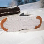 A close-up of a shoe sole resting in snow, showing a white treaded bottom with tan rubber protective corners.