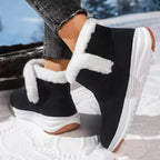 A person wearing black winter ankle boots with thick white faux-fur trim and chunky white soles, standing outdoors on snow.