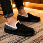 Black slippers with white and gray fur lining worn indoors on a patterned rug.
