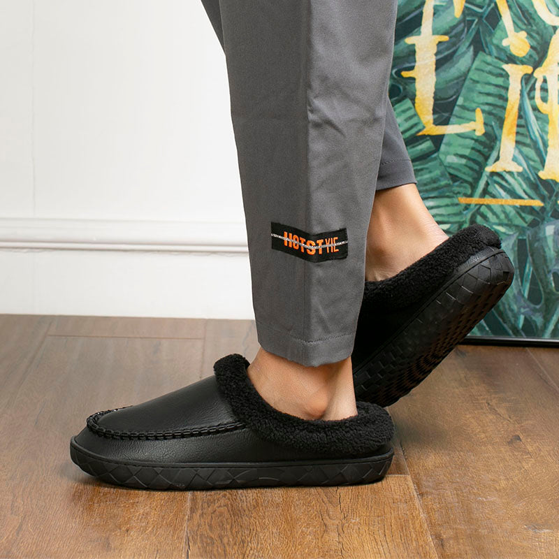 Person wearing black slippers with a brand logo on a wooden floor.
