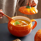 Orange pumpkin-themed mug with soup and a spoon, held by a person.